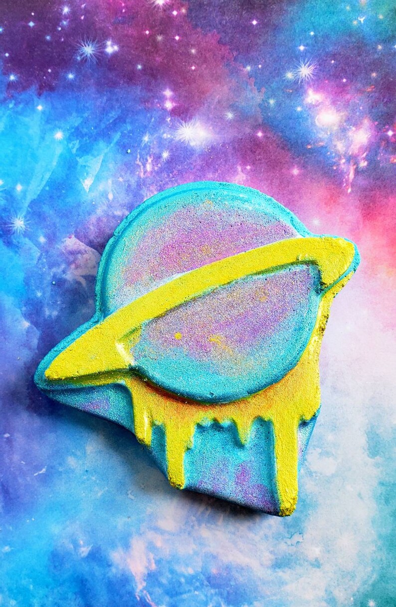 May include: A colorful bath bomb shaped like a planet with a yellow ring and dripping accents. The planet is a mix of blue, purple, and pink with a yellow ring. The background is a vibrant galaxy of blues, purples, and pinks.