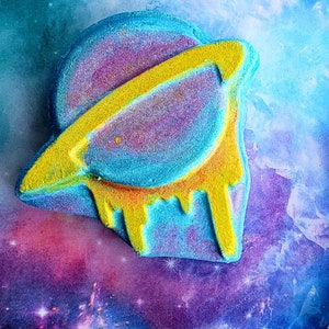 May include: A colorful bath bomb shaped like a planet with a yellow ring and dripping yellow accents. The planet is a mix of blue, purple, and yellow, set against a galaxy-themed background. This unique bath bomb adds a touch of cosmic fun to bath time.