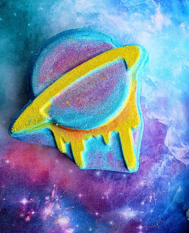 May include: A colorful bath bomb shaped like a planet with a yellow ring and dripping yellow accents. The planet is a mix of blue, purple, and yellow, set against a galaxy-themed background. This unique bath bomb adds a touch of cosmic fun to bath time.
