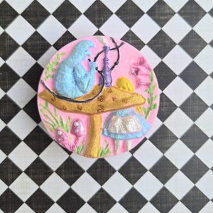 May include: A pink, round decorative piece featuring a blue figure with a hookah, seated at a table atop a mushroom. The mushroom has a gold top and a blue and silver skirt. The background includes pink, green, and purple elements.