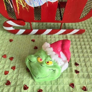 May include: A green Grinch-shaped bath bomb with a red and white Santa hat, placed on a green textured surface. A red sleigh and candy cane are in the background. Red heart-shaped confetti is scattered around the bath bomb.