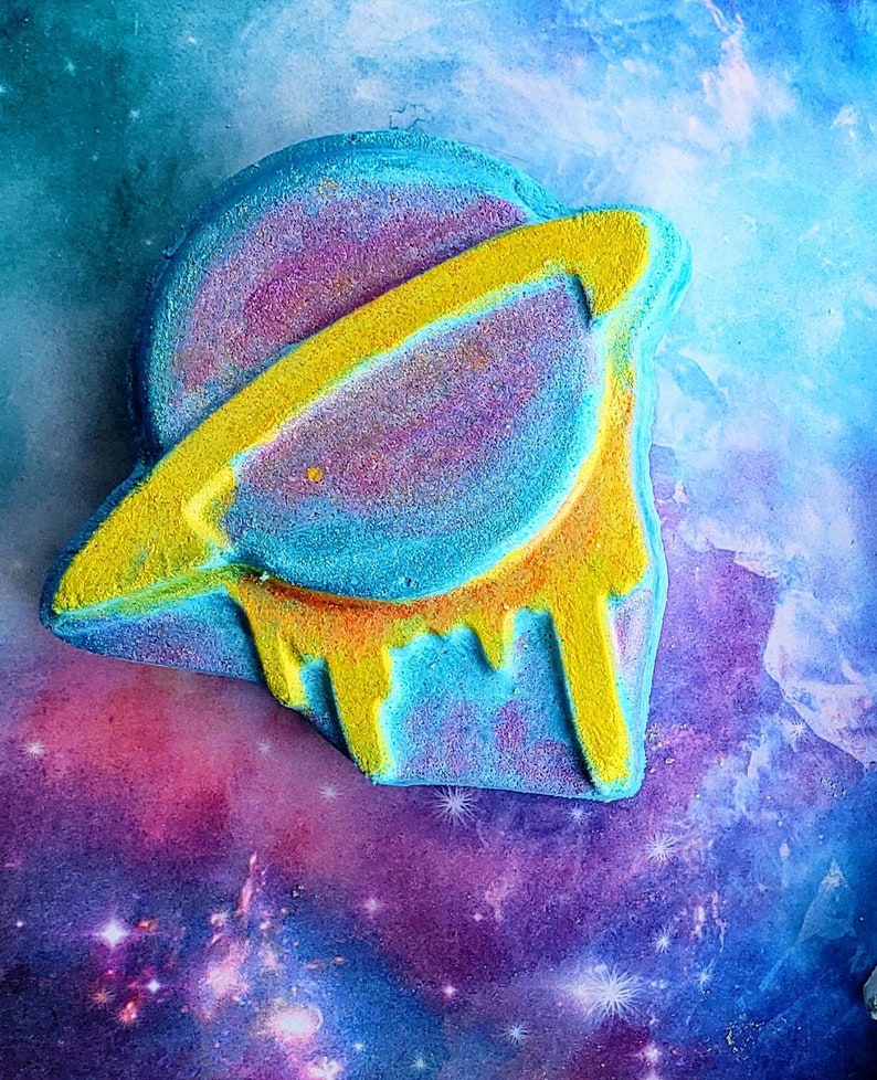 May include: A colorful bath bomb shaped like a planet with a yellow ring. The planet is a mix of blue, purple, and yellow, with a dripping yellow design on the bottom. The background is a galaxy of blues, purples, and white.