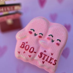 May include: A pink, heart-shaped decoration with two ghost figures hugging. The ghosts have black eyes and rosy cheeks. The words "BOO-STIES" are written in dark red. The background is a light purple with heart shapes.