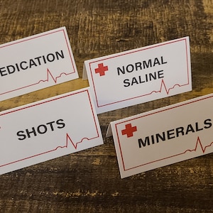May include: Four white table cards with red borders and a red cross. The cards have a red heartbeat graphic and the text "MEDICATION", "NORMAL SALINE", "SHOTS", and "MINERALS".
