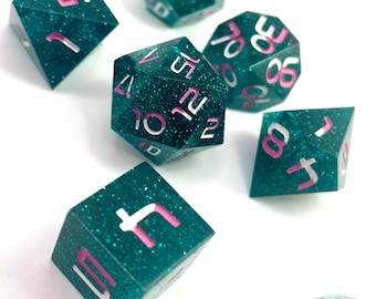 Può includere: Un set di sette dadi poliedrici color teal con numeri rosa e bianchi. I dadi sono sparsi su una superficie bianca, mostrando le loro varie forme geometriche, tra cui un d4, d6, d8, d10, d12 e d20.