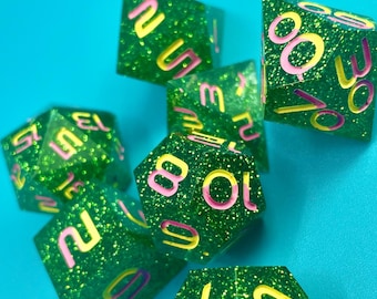 Può includere: Un gruppo di sette dadi poliedrici verdi e glitterati con numeri rosa e gialli. I dadi sono sparsi su una superficie blu brillante, evidenziando il loro design sfaccettato e la vivace combinazione di colori. Ideale per i giochi da tavolo.