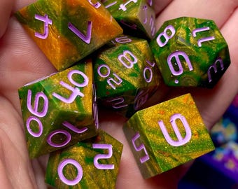 Può includere: Un set di sette dadi poliedrici di varie forme, con motivi verdi e dorati e numeri viola. I dadi sono tenuti in una mano. Ogni dado è progettato per i giochi di ruolo.