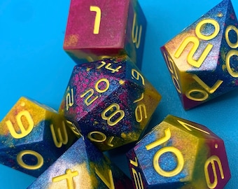 May include: A set of seven polyhedral dice with a galaxy-inspired design. The dice are made of resin and have a deep blue, purple, and gold colour scheme with yellow numbers.