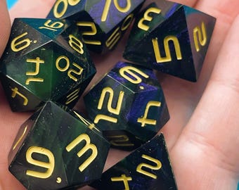 May include: A set of seven polyhedral dice in various shapes, held in a hand. The dice are black with gold numbering and swirls of green and purple. The dice are for tabletop games.