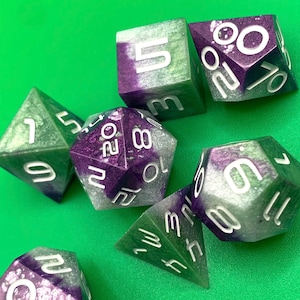 May include: Set of six green and purple dice with white numbers. The dice are made of a translucent material with a glittery finish.