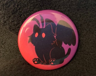 May include: A pink and purple button with a black silhouette of a cat with wings and horns. The cat has large, white eyes.