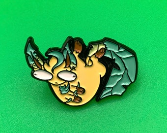 May include: A yellow and green enamel pin of a cartoon creature with large eyes and a curled tail. The creature is holding its wings in front of its face.