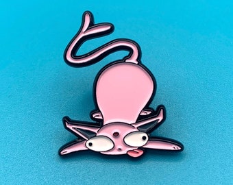 May include: A pink enamel pin depicting a cartoon cat in a comical pose. The cat has large eyes, a protruding tongue, and a long, curved tail. The pin has black outlines and is set against a blue backdrop.