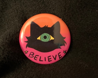 May include: A pink and orange button featuring a black cat silhouette with a green eye. The cat has a yellow eyelid and the button reads "BELIEVE" in black lettering.