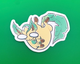 May include: A die-cut sticker featuring a cartoon character with a light yellow body, brown paws and ears, and green leaf-like accents. The character has large, blank eyes and is lying on its back. The sticker is outlined in black.