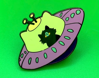 May include: A purple and green enamel pin of a black cat in a flying saucer. The cat has one large green eye.