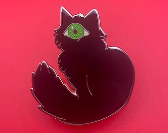 May include: A black cat enamel pin with a large green eye. The cat is sitting with its tail curled around its body. The pin has a silver outline and is set against a red background.