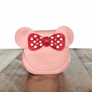 May include: A pink faux leather purse with a red bow with white polka dots. The purse has two ears on the top, resembling a mouse.