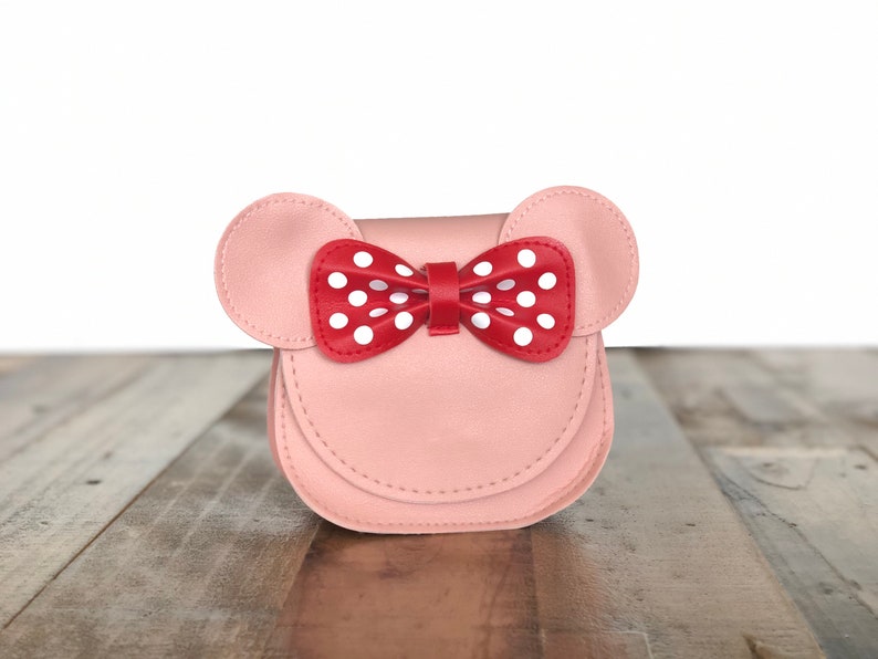 May include: A pink faux leather purse with a red bow with white polka dots. The purse has two ears on the top, resembling a mouse.