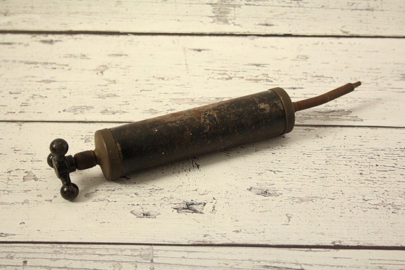 May include: A vintage metal hand pump with a black finish and a long, thin nozzle. The pump has a T-shaped handle with three round knobs.