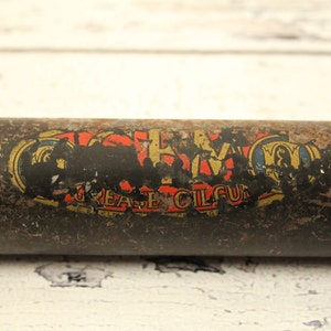 May include: A vintage metal container with a faded label that reads "Grease Oil Cup". The container is rusty and has a black background with gold and red lettering.