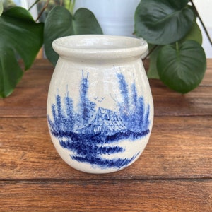 May include: A white ceramic vase with a blue and white landscape design. The vase is round and has a wide mouth.
