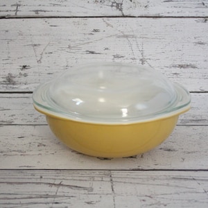 May include: A yellow glass baking dish with a clear glass lid. The dish is sitting on a white wooden surface.