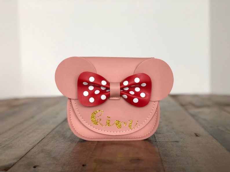 May include: A pink faux leather purse with a red and white polka dot bow and the name "Kimi" written in gold glitter.