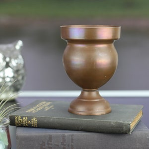 May include: A copper-coloured vase with a rounded body and a flared lip sits on top of two books. The vase is on a dark surface.