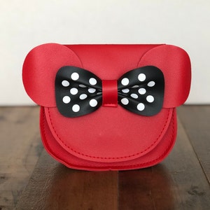 May include: A red faux leather purse with a black and white polka dot bow. The purse has two rounded ears on the top.