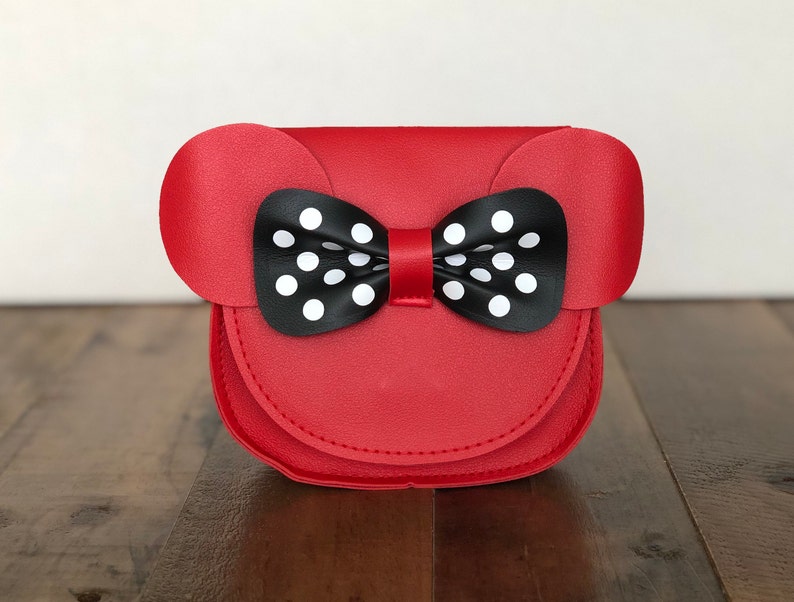 May include: A red faux leather purse with a black and white polka dot bow. The purse has two rounded ears on the top.