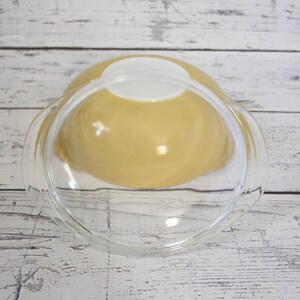 May include: A yellow glass mixing bowl with a clear glass lid. The bowl is sitting on a white wooden surface.