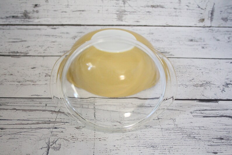 May include: A yellow glass mixing bowl with a clear glass lid. The bowl is sitting on a white wooden surface.