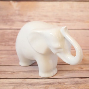 Pode incluir: Uma figura de elefante de cerâmica branca com a tromba levantada. O elefante está em pé sobre uma superfície de madeira.