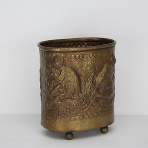 May include: A brass planter with an oval shape and an embossed design of monkeys and leaves. The planter has three small feet and a slightly flared top.