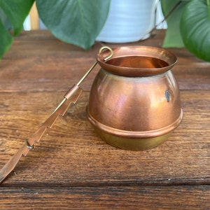 Puede incluir: Una tetera de cobre y latón con un asa larga y fina. La tetera tiene un cuerpo redondo y un pequeño pico. El asa está unida a la tetera con un pequeño lazo.