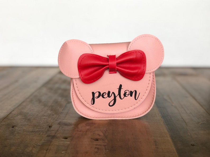 May include: A pink faux leather purse with a red bow and the name "peyton" written in black.