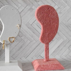 May include: Two earring display stands, one white and one pink. The white stand has a gold cross earring, a gold heart earring, and a gold hoop earring. The pink stand is made of velvet.