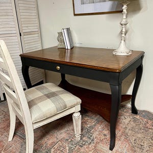 May include: A wooden desk with a dark brown top and black legs featuring a single drawer with a gold knob. Beside it sits a white chair with a beige and gray plaid upholstered seat. The desk has a lower shelf and curved legs.