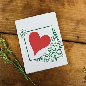 May include: A white card with a green frame and a red heart inside. The frame is decorated with white flowers and green leaves.