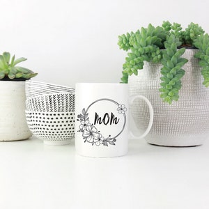 May include: White ceramic mug with a black floral design and the word "MOM" in a circle. The mug is sitting on a white surface with two small white bowls and a potted succulent plant.