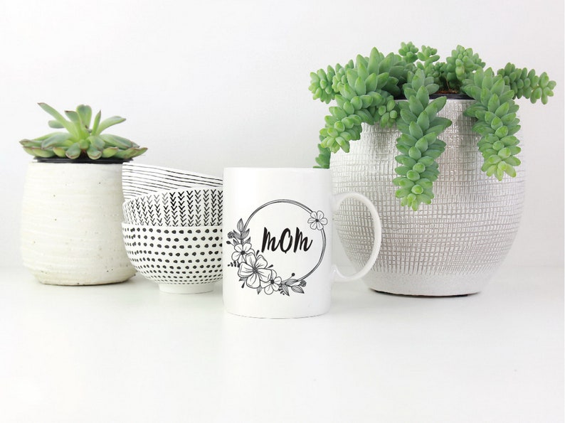 May include: White ceramic mug with a black floral design and the word "MOM" in a circle. The mug is sitting on a white surface with two small white bowls and a potted succulent plant.