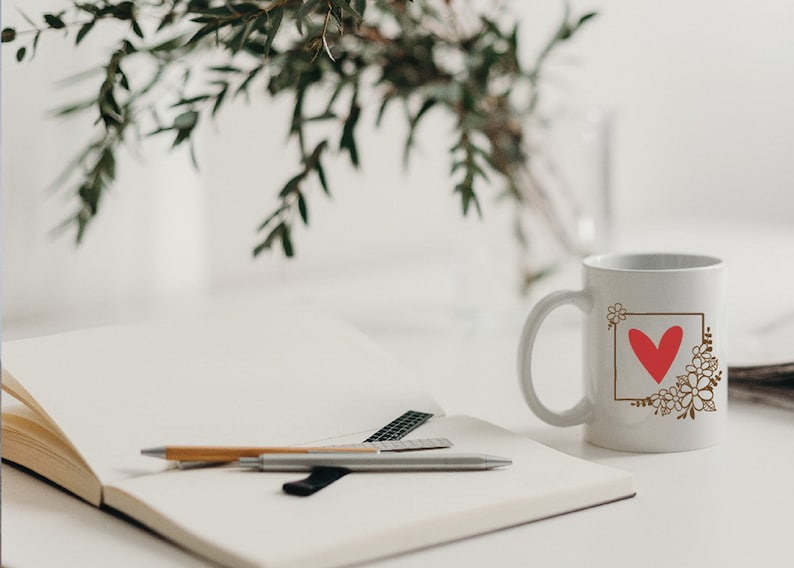 May include: White ceramic coffee mug with a brown floral design and a red heart in a square frame. The mug is sitting on a white table next to a notebook and pens.