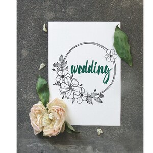 May include: A white card with a black floral wreath and the word "wedding" written in teal green.