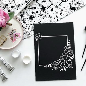 May include: A black card with a white floral border design. The border is a square frame with a corner filled with white flowers and leaves. The card is surrounded by calligraphy pens and paint tubes.
