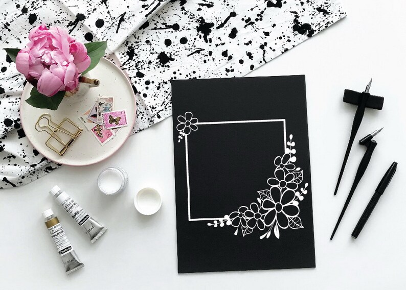 May include: A black card with a white floral border design. The border is a square frame with a corner filled with white flowers and leaves. The card is surrounded by calligraphy pens and paint tubes.