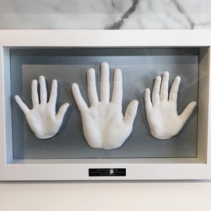 May include: White framed shadow box containing three white handprints. The handprints are arranged horizontally, with the largest handprint in the centre. The shadow box has a silver background and a small black plaque with text at the bottom.