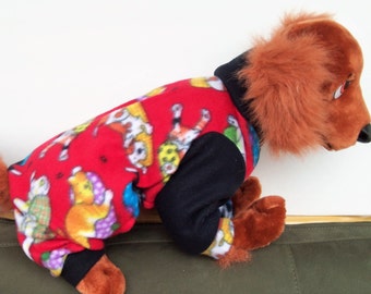 May include: A red, black, and white dog pajamas with a cat print. The pajamas have a black hood and a black sleeve. The dog is wearing the pajamas and is sitting on a green surface.