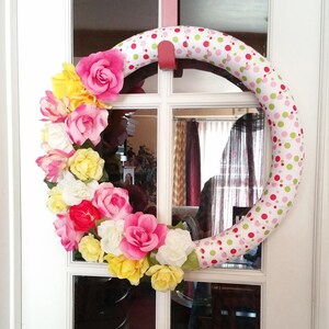 Puede incluir: Una corona de lunares rosa, blanca y verde con un arreglo floral de flores rosas, amarillas y blancas. La corona está colgada en una puerta blanca.
