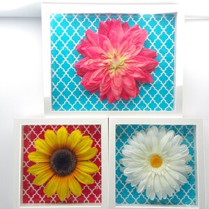 Puede incluir: Tres impresiones florales enmarcadas con marcos blancos. La impresión superior tiene una flor rosa sobre un fondo turquesa. La impresión del medio tiene un girasol amarillo sobre un fondo rojo. La impresión inferior tiene una margarita blanca sobre un fondo turquesa.
